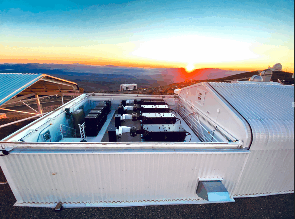 The shed-like enclosure of LVM with the roof rolled off. Inside are four small telescopes with black boxes next to them.